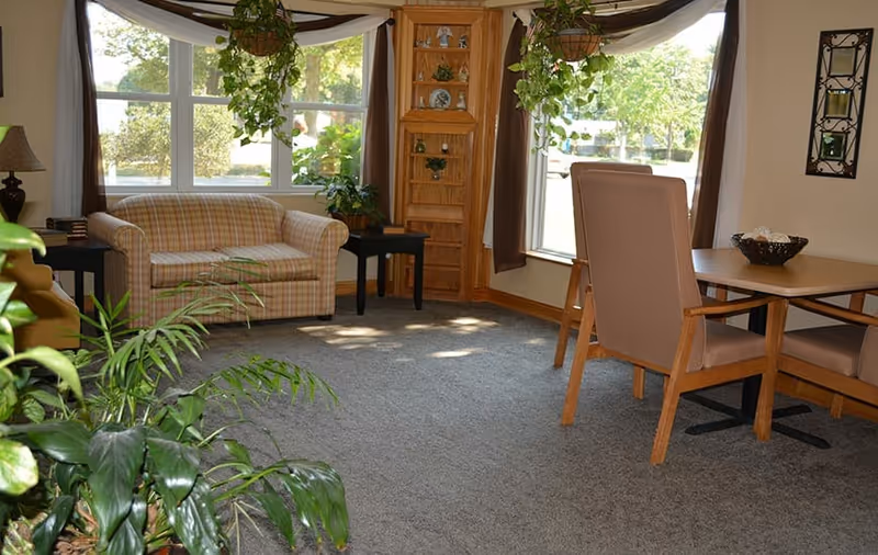 A cozy living room area with a plaid loveseat, two side tables with lamps, a wooden corner shelf with decorative items, and a dining table with four chairs. Large windows with brown curtains let in natural light, and several green plants add a touch of nature to the room.