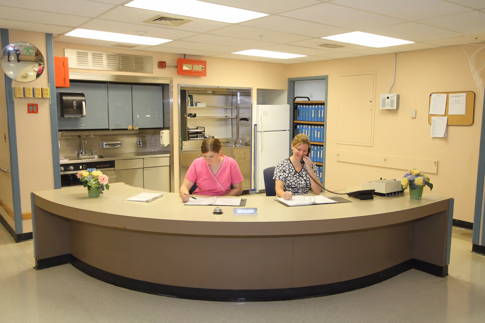 Two staff members sit at a curved reception desk in a facility's reception/nurses' station, one speaking on the phone and one writing.