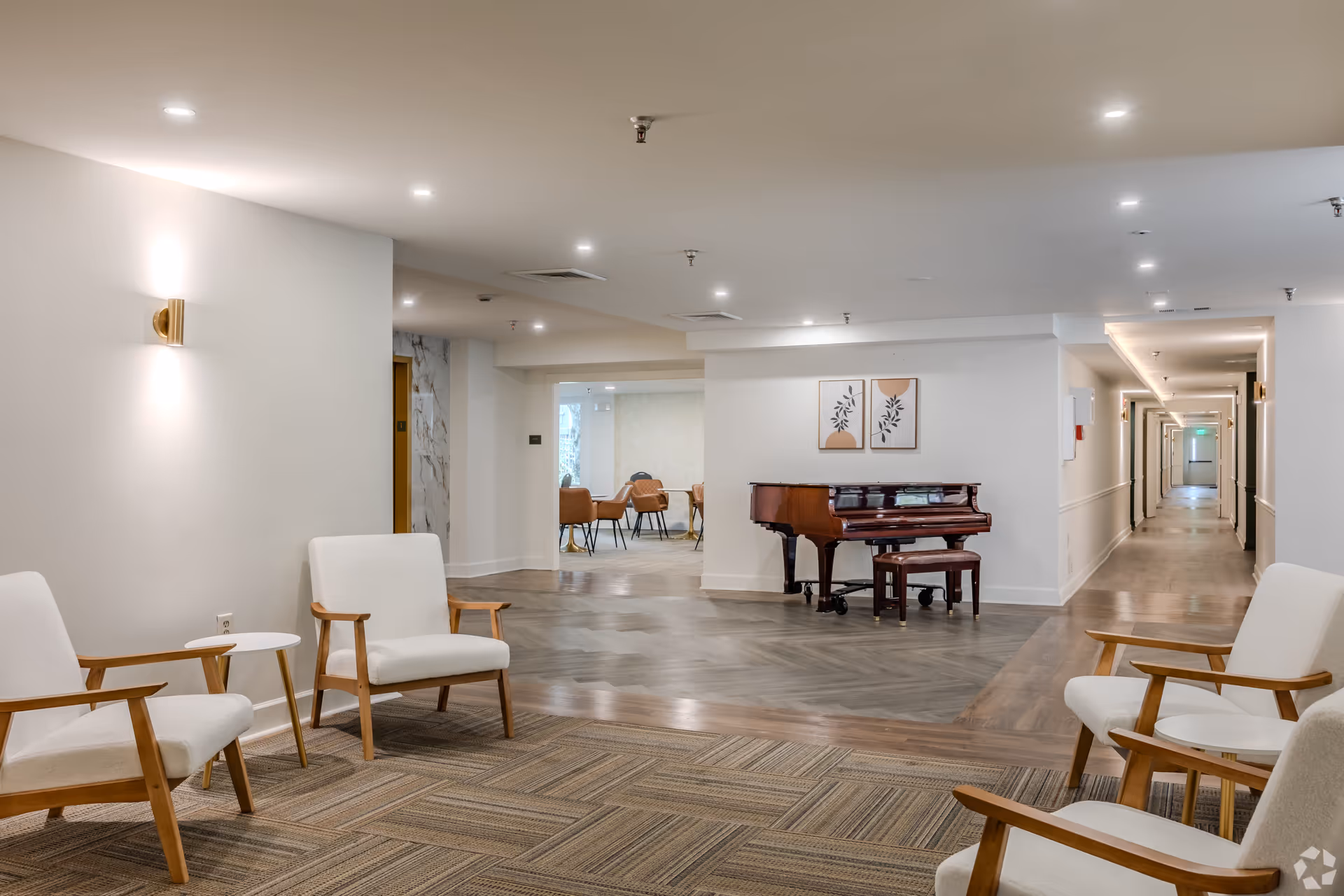 A spacious and well-lit interior common area in a senior living facility featuring four white cushioned chairs with wooden armrests arranged around a small white side table. In the background, there is a polished wooden piano with a matching bench, two framed abstract art pieces on the wall above it, and a hallway leading to other rooms. The flooring transitions from carpet to wood, and recessed ceiling lights illuminate the space.