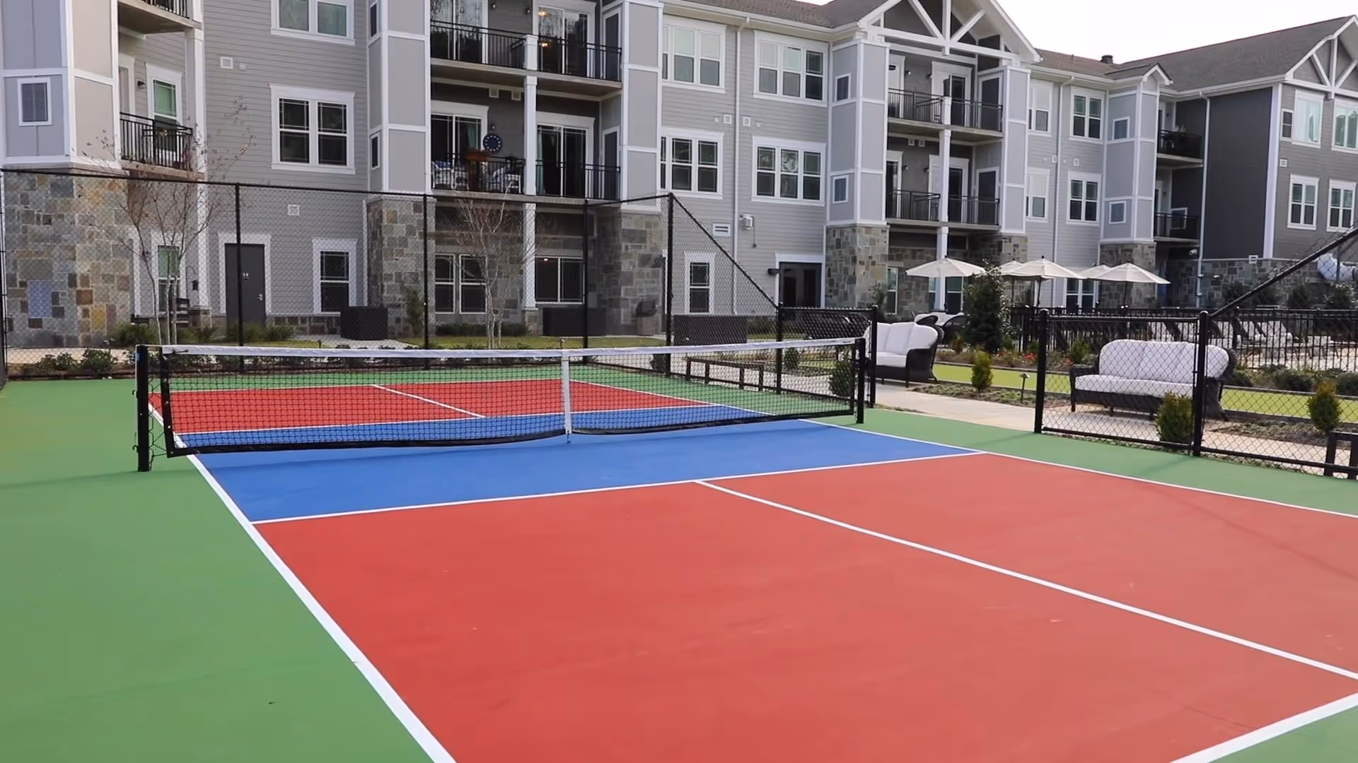 Outdoor tennis or pickleball court with red, blue, and green surface colors, surrounded by a black chain-link fence. In the background, there is a multi-story residential building with balconies and stone accents. Patio furniture with umbrellas is visible near the court.