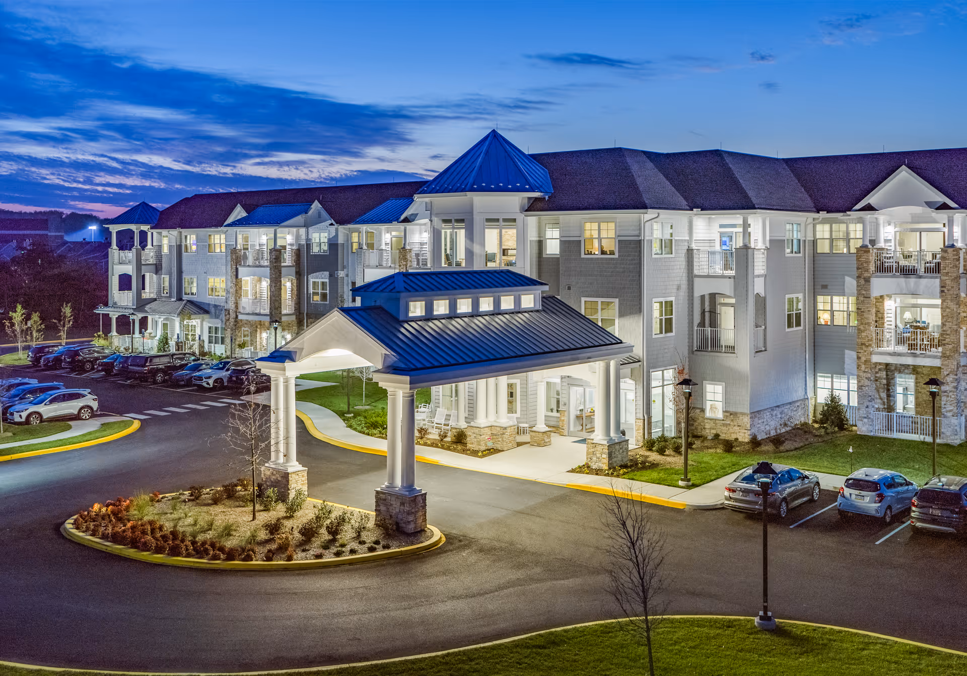 Exterior view of The Lodge at Historic Lewes senior living facility at dusk, showing a large, well-lit building with multiple floors, balconies, a covered entrance, and a parking lot with several cars.