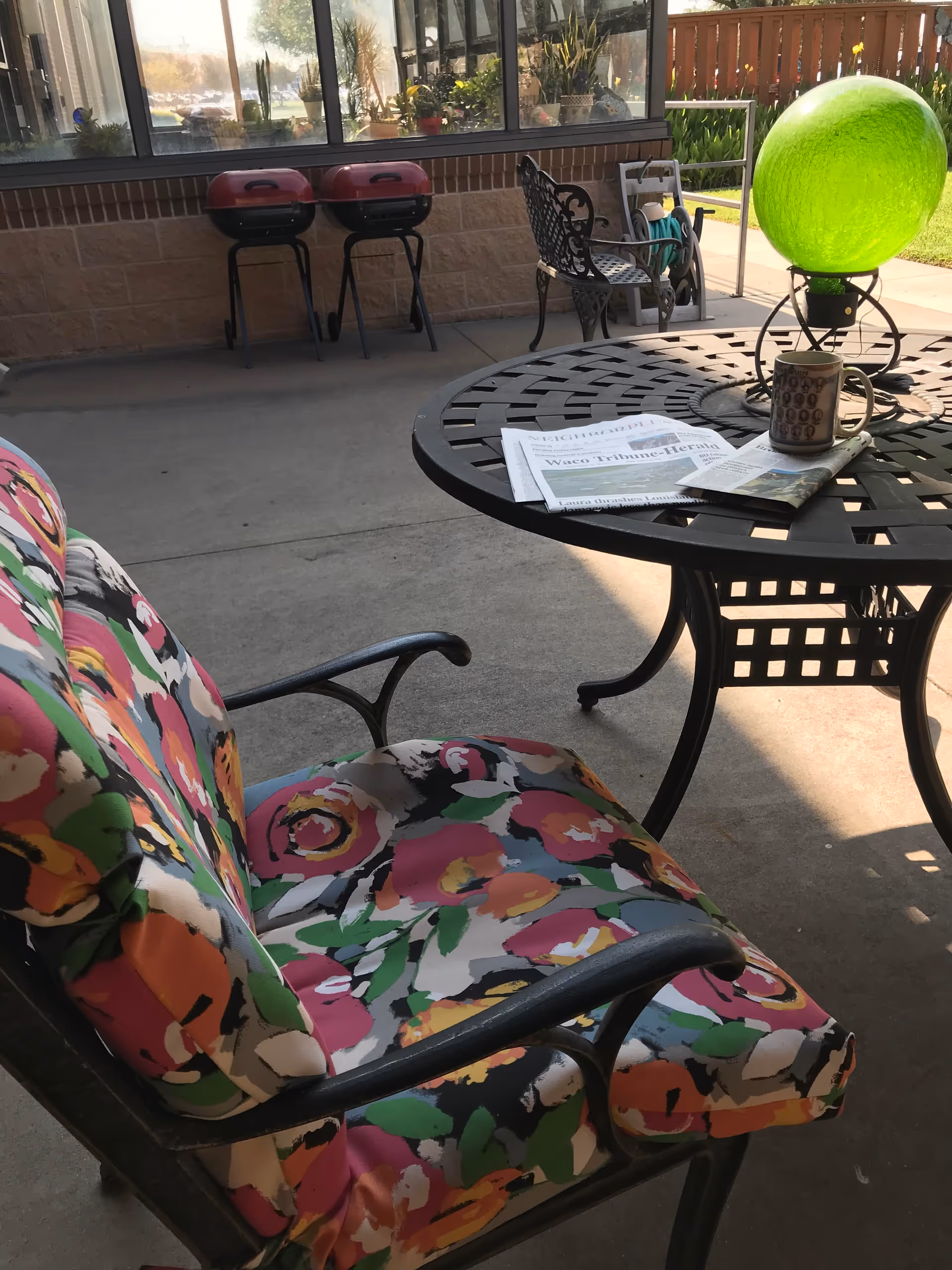 Patio seating area with a floral-cushioned chair, round metal table holding a newspaper and mug, and grills in the background.