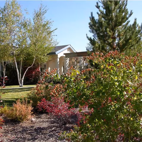 Landscaped front yard with trees and flowering shrubs in front of a beige memory care building under a clear blue sky.