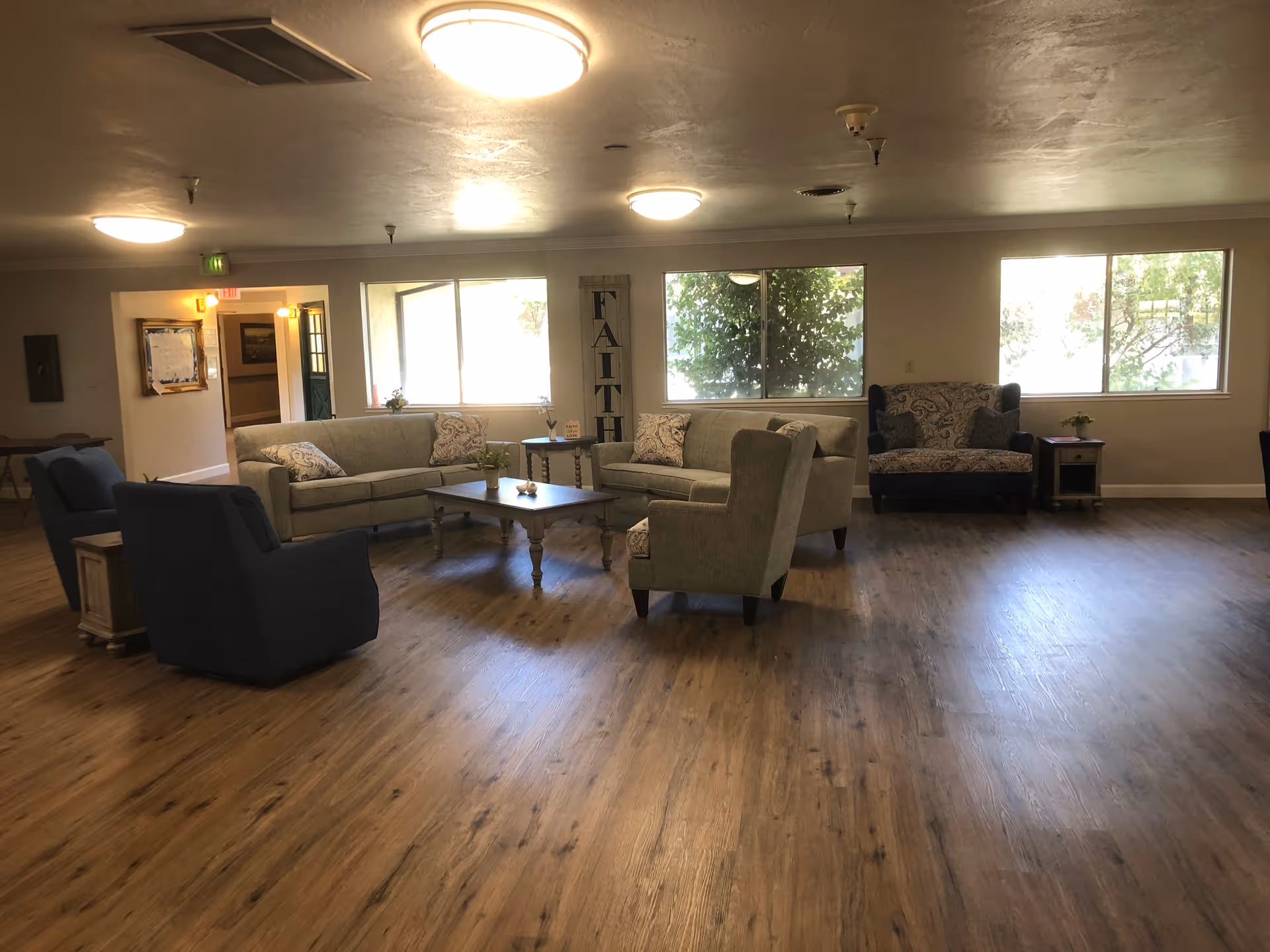 A spacious living room area with wooden flooring, featuring two beige sofas, two armchairs, a coffee table, and side tables. Large windows allow natural light to brighten the room. A decorative sign with the word 'FAITH' is displayed between the windows.