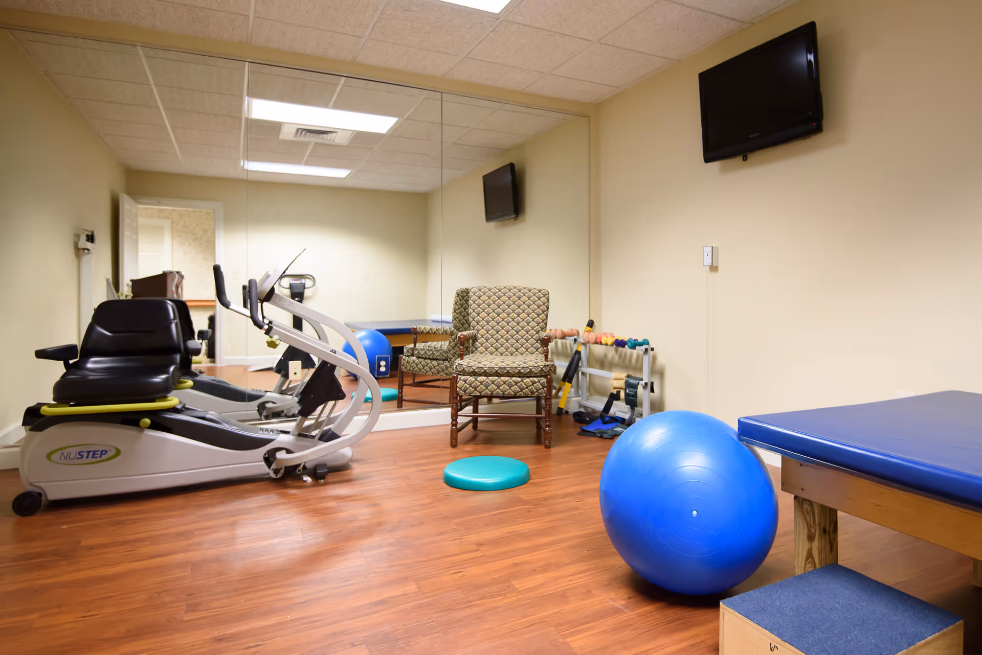 A small exercise room with a recumbent exercise bike, a large blue exercise ball, a padded exercise table, two patterned armchairs, a set of small dumbbells on a rack, and a large wall mirror reflecting the room. A flat screen TV is mounted on the wall above the exercise table.