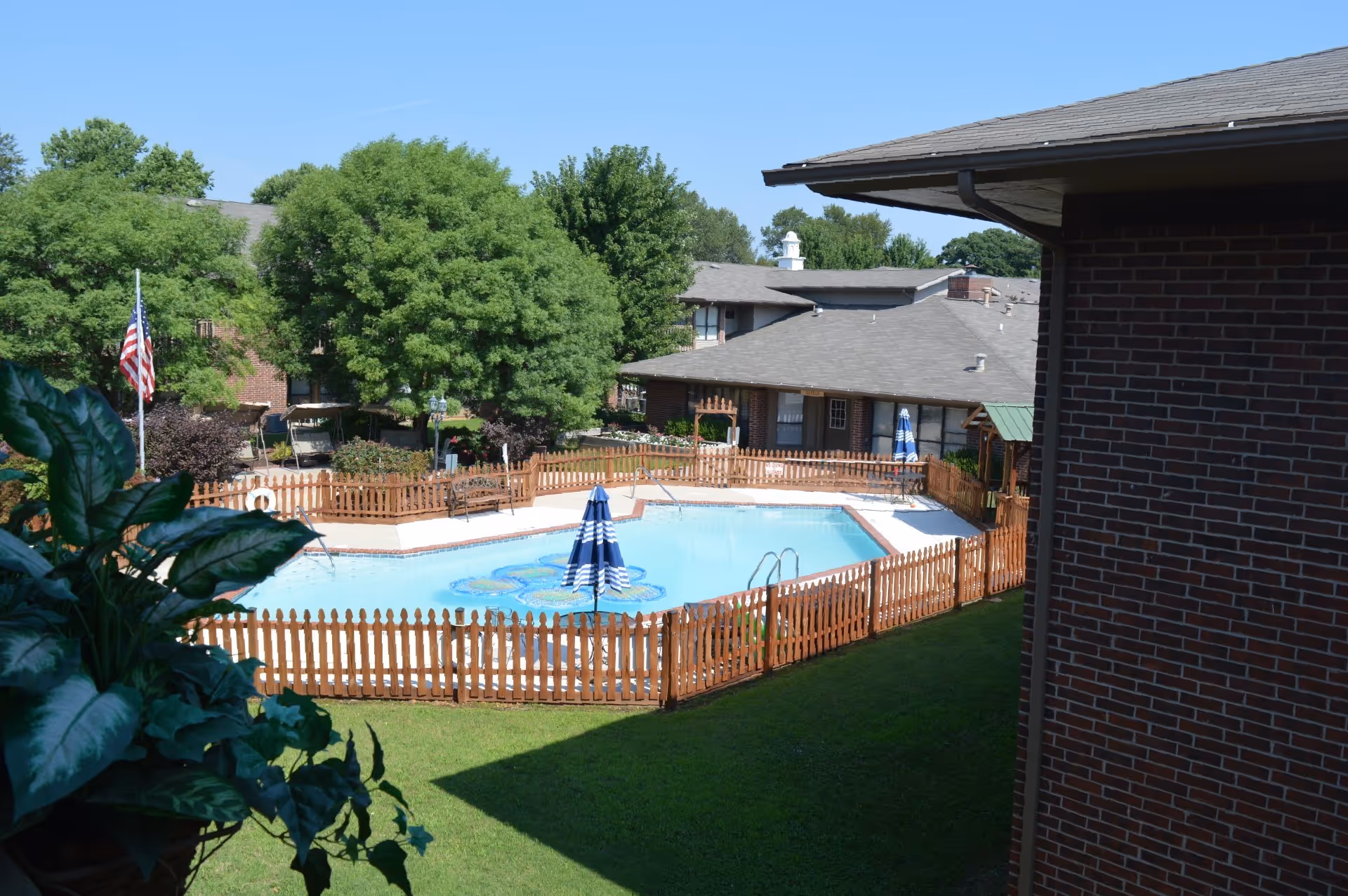 Fenced outdoor swimming pool in a landscaped courtyard surrounded by brick senior living buildings.