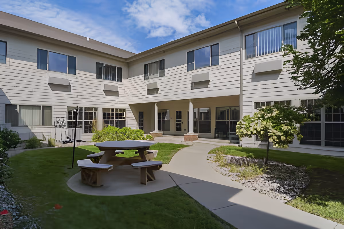 Outdoor courtyard area of a senior living facility with a round picnic table and benches on a concrete pad, surrounded by green grass, shrubs, and a small tree. The building is two stories with multiple windows and a covered walkway supported by columns. The sky is partly cloudy.