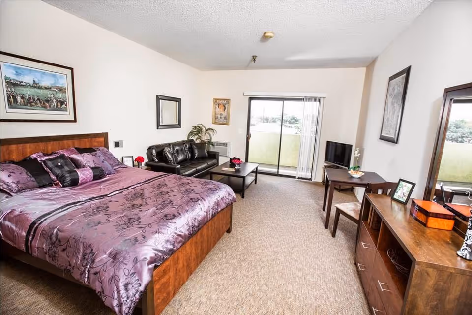 A spacious senior living bedroom featuring a large bed with purple floral bedding, a black leather couch, a coffee table, a small dining table with two chairs, a flat-screen TV on a stand, and a wooden dresser with a mirror. The room has beige carpet, white walls with framed artwork, and a sliding glass door leading to a balcony with a view outside.