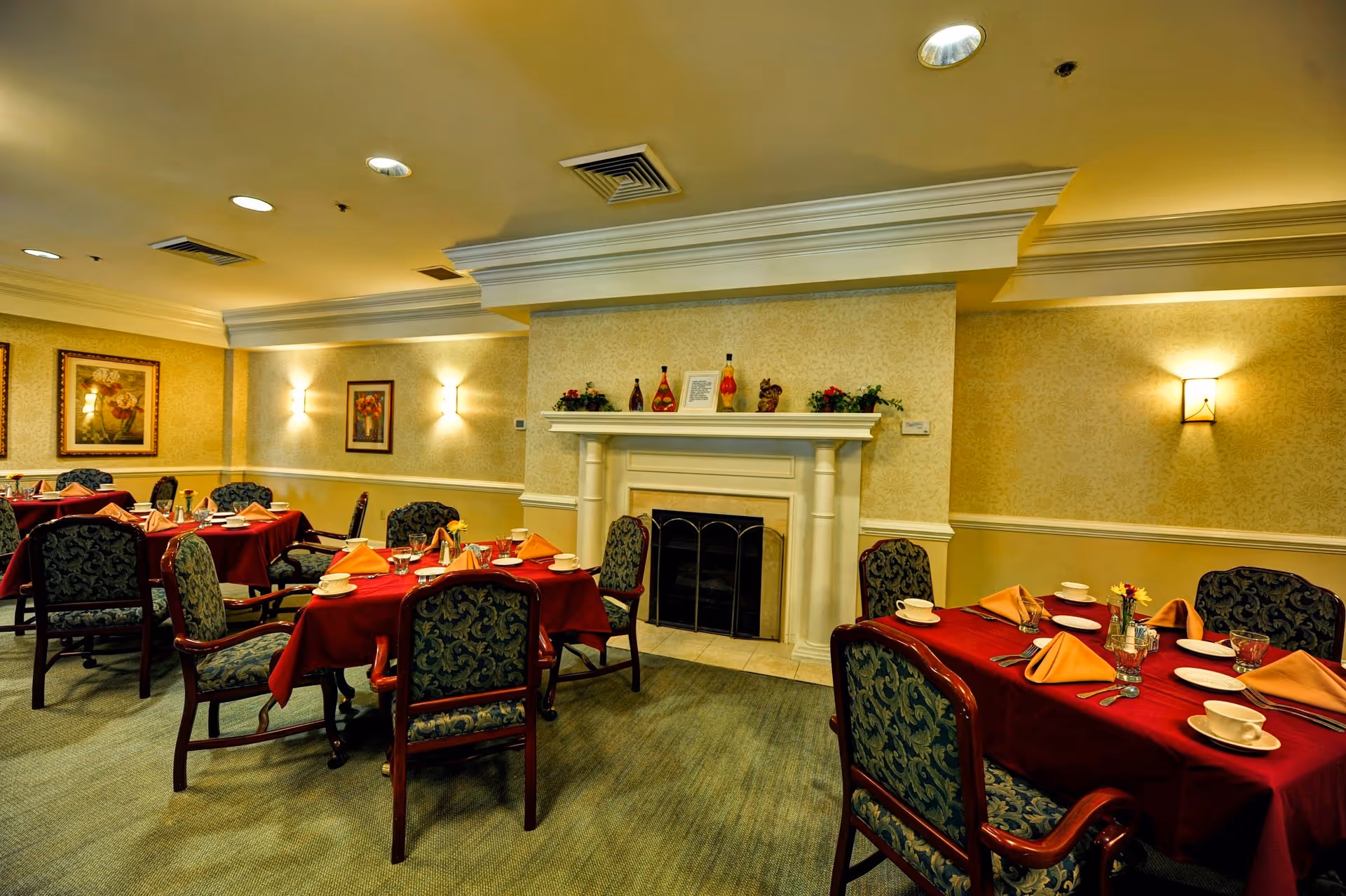 A dining room with several tables covered in red tablecloths and set with plates, cups, utensils, and folded orange napkins. The room has patterned upholstered chairs, a fireplace with decorative items on the mantel, framed artwork on the walls, and warm lighting from wall sconces and ceiling lights.