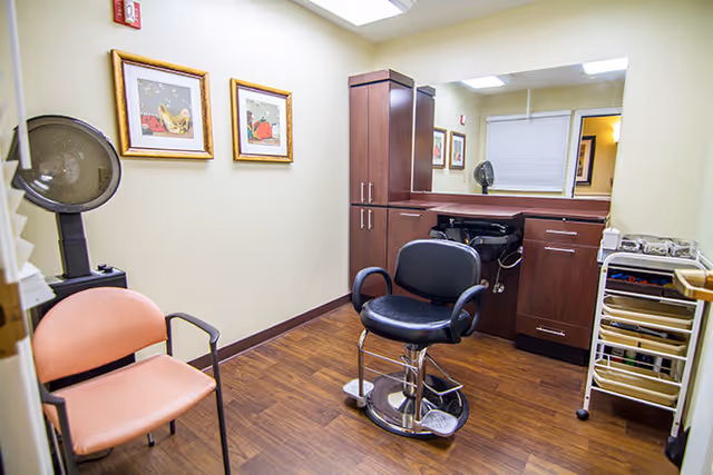 A small salon or grooming room with a black salon chair in front of a counter with a sink and cabinets. There is a peach-colored waiting chair, a hair dryer, and a rolling cart with supplies. The walls are light yellow with two framed pictures hanging.