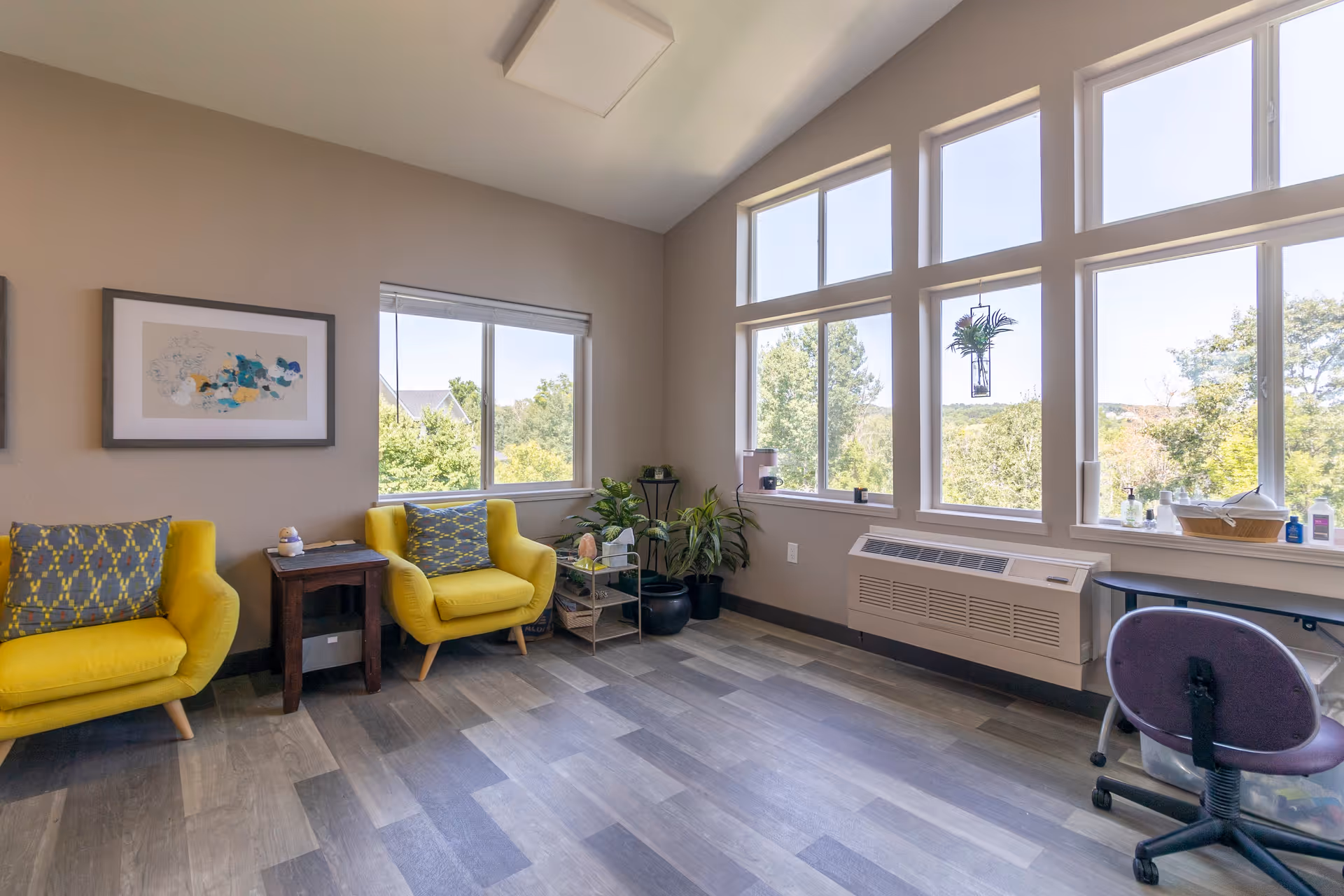 A bright room with large windows showing a view of trees outside. Inside, there are two yellow armchairs with patterned cushions, a small wooden side table between them, several potted plants, and a purple office chair near a window ledge with various small items on it. The floor has a wood-like pattern, and the walls are painted beige.