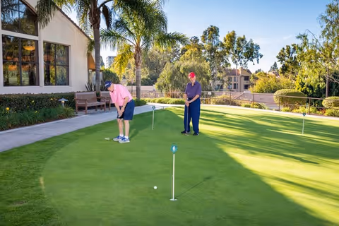 Two elderly men playing golf on a putting green surrounded by palm trees and bushes, with a building and benches visible in the background under a clear blue sky.