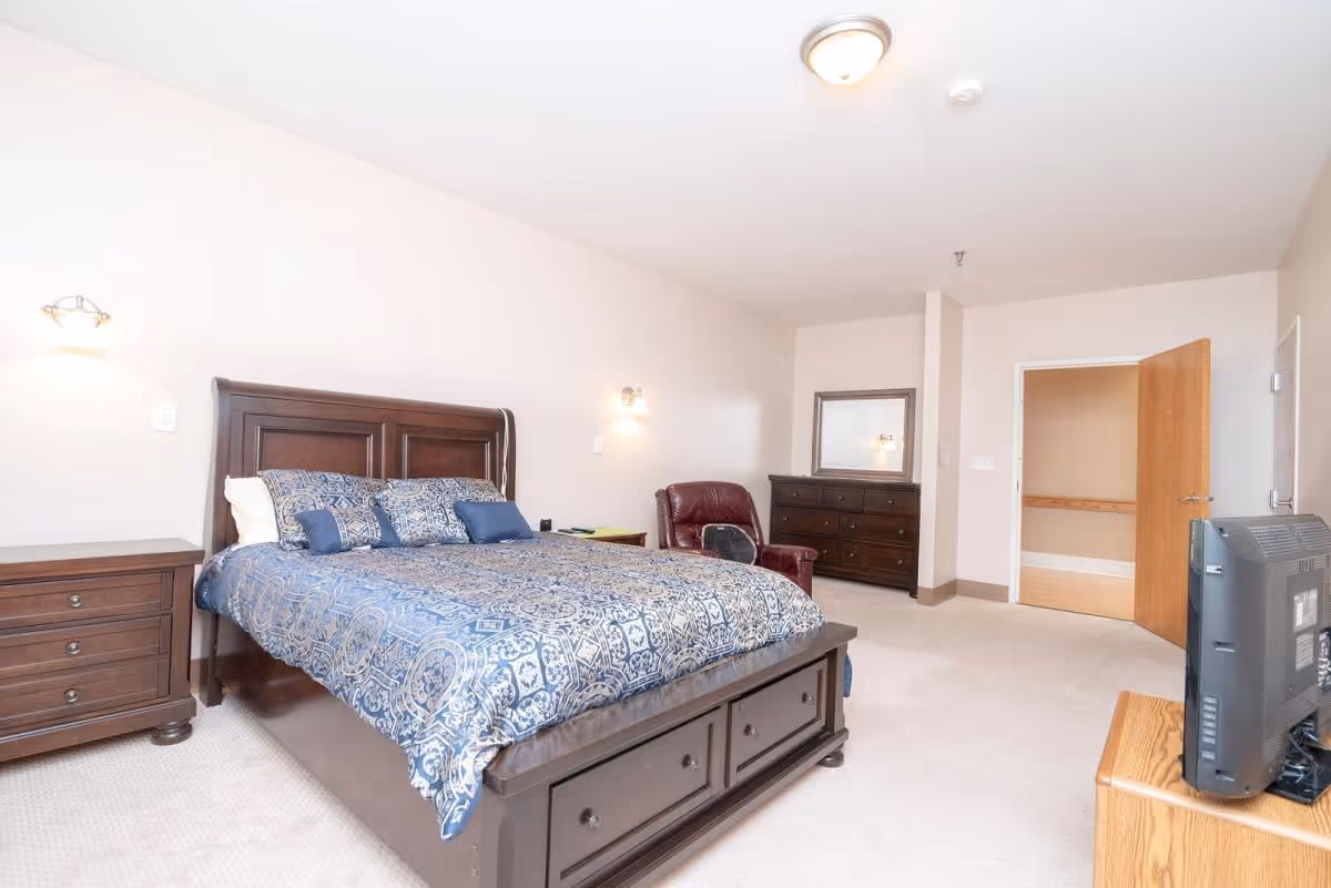 A spacious bedroom with a large wooden bed featuring a blue patterned bedspread and multiple pillows. There is a matching wooden nightstand with drawers on the left side of the bed, a dark red leather armchair, a wooden dresser with a mirror, and a television on a wooden stand. The room has beige walls and carpeted flooring, with a door open to an adjacent hallway.