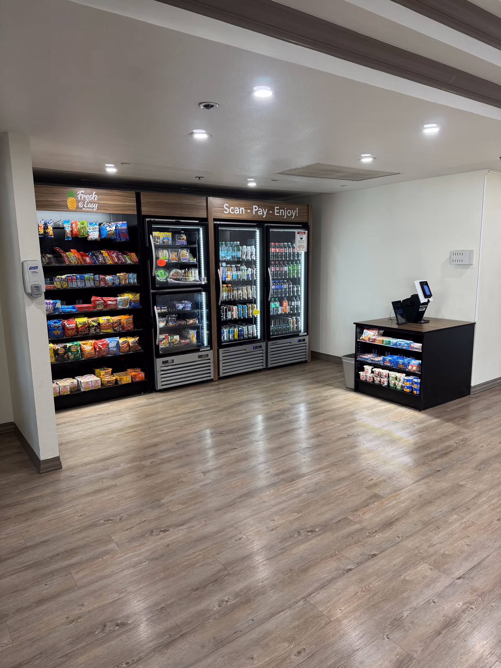 A small convenience area inside a facility with shelves stocked with snacks and refrigerated coolers containing drinks and packaged food. There is a self-checkout kiosk on a counter to the right. The floor is wood-style laminate and the walls are white.