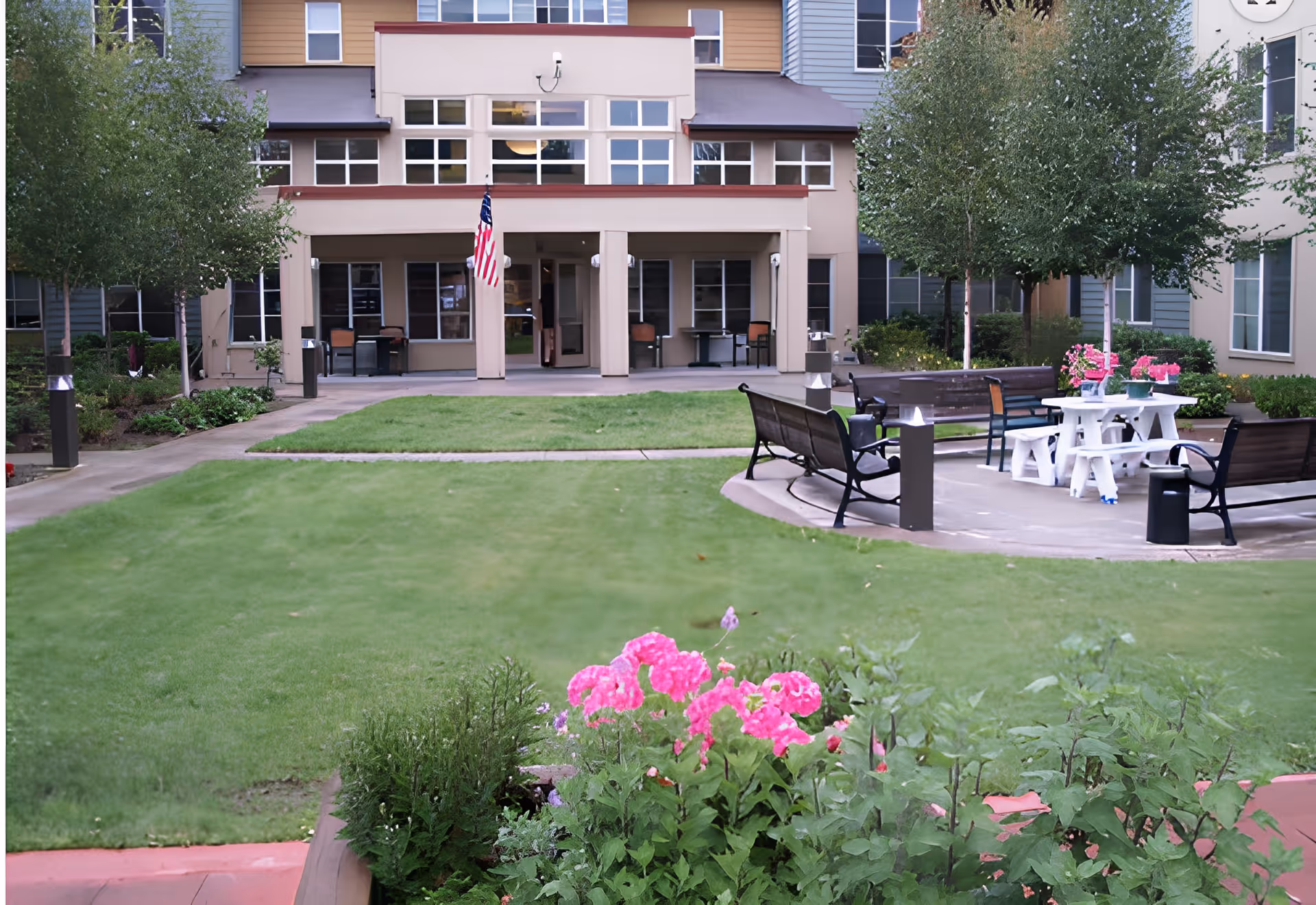 Outdoor courtyard area of a senior living facility with green grass, flower beds with pink flowers, benches arranged around a white picnic table, and trees. The building in the background has multiple windows and an American flag hanging near the entrance.