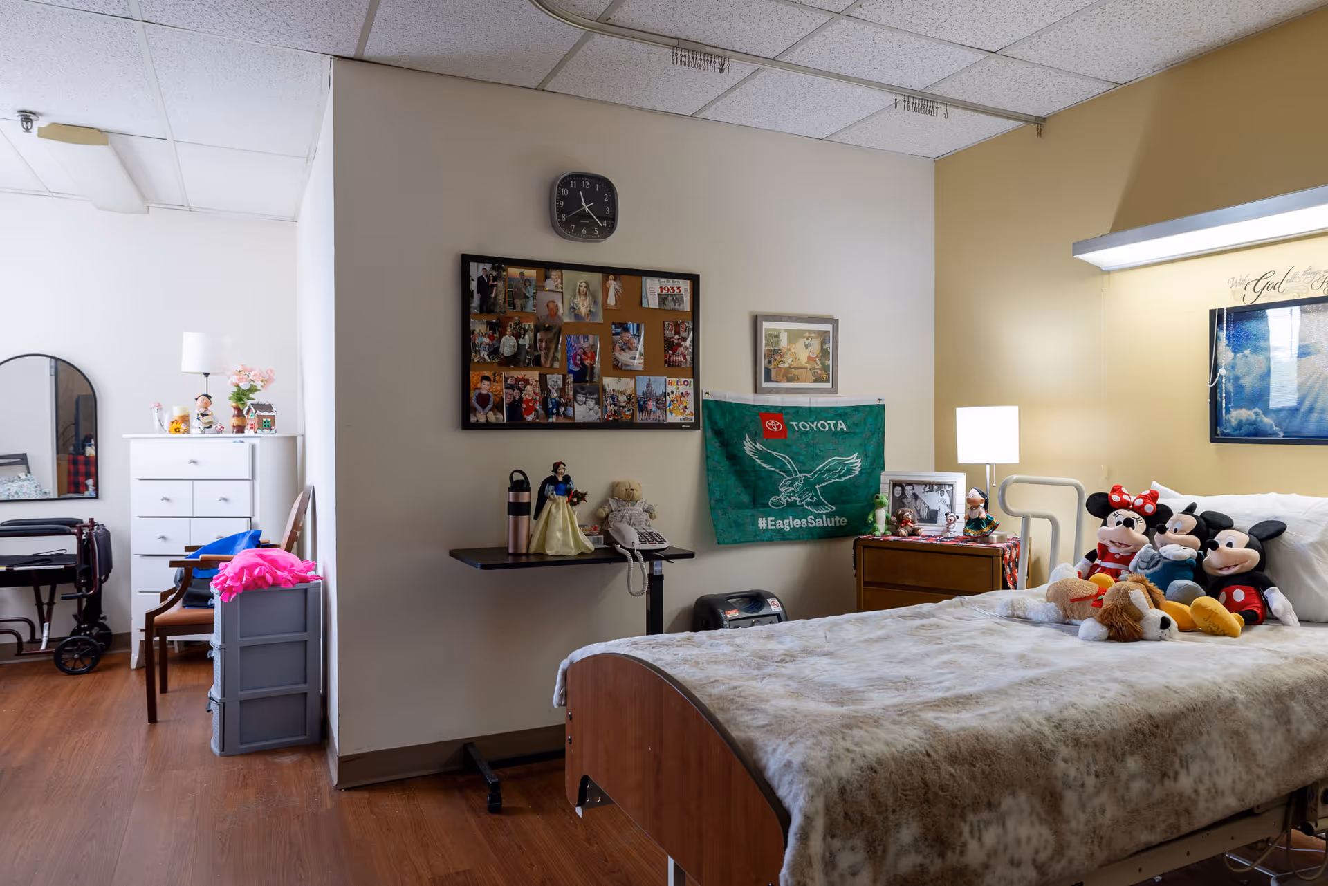 A cozy bedroom in a senior living facility featuring a single bed with a beige blanket and several stuffed toys including Mickey and Minnie Mouse. The room has a wooden floor, a nightstand with a lamp, a bulletin board with photos, a green Toyota Eagles Salute flag on the wall, and a dresser with a mirror and flowers. There is also a walker and a chair near the dresser.