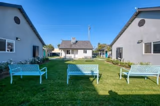 Green grassy courtyard flanked by single-story buildings with three turquoise benches facing a small house under a clear blue sky.