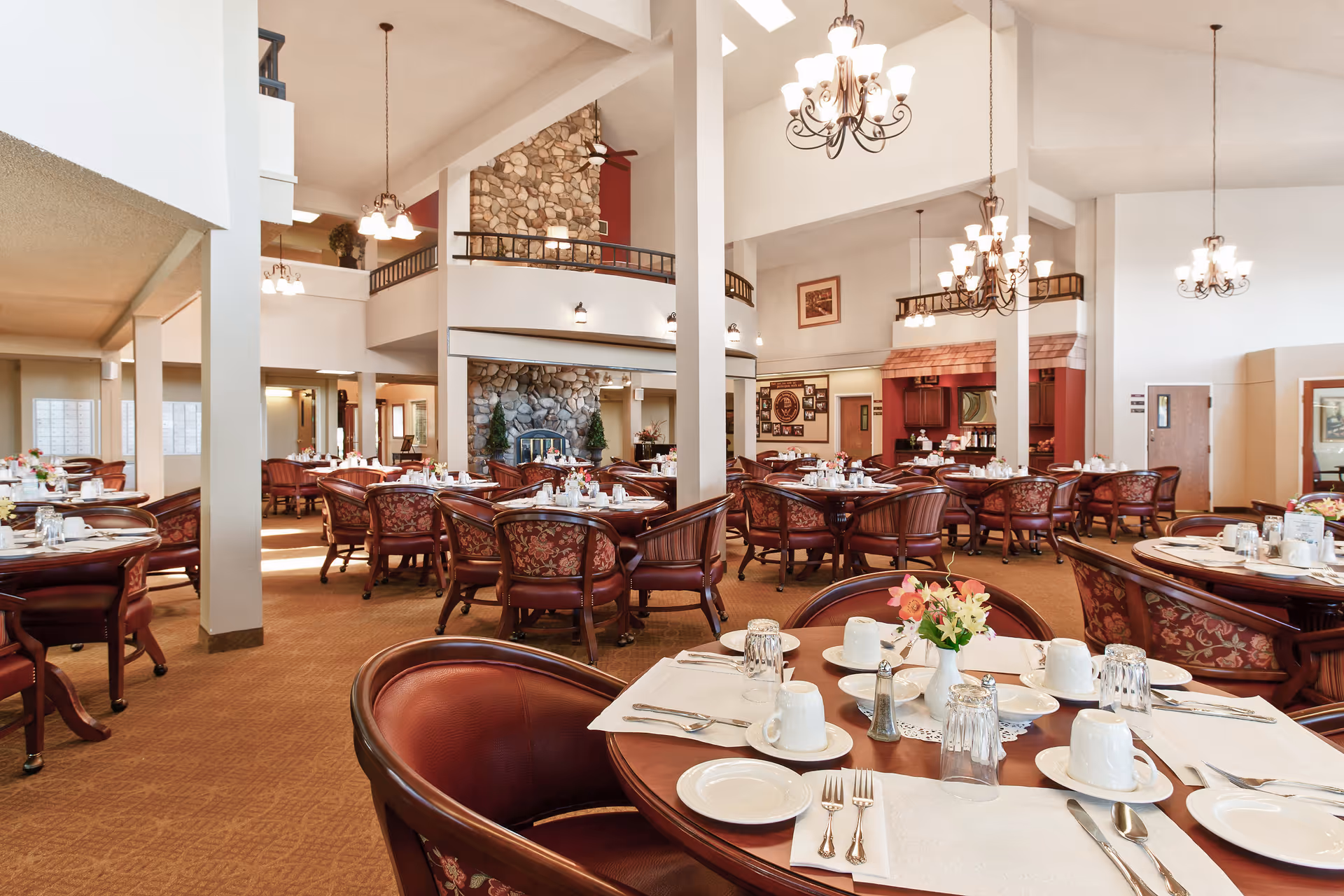 Large airy dining room with many round tables set for service, upholstered chairs, chandeliers, and a stone fireplace.
