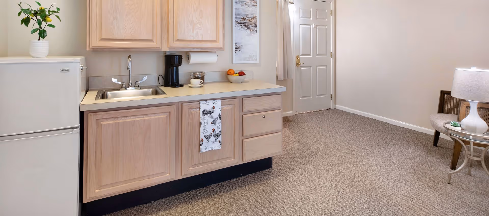 A small kitchenette with light wood cabinets, a sink, mini fridge and coffee maker opening into a carpeted living area with a door and side table lamp.