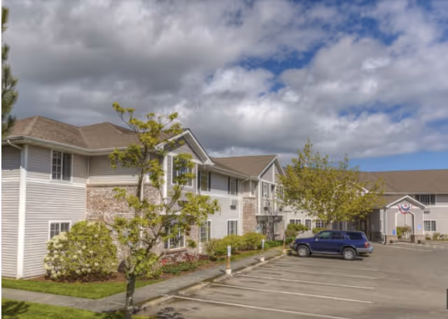 Exterior view of a two-story senior living facility with parking lot, landscaping, and a parked SUV under a partly cloudy sky.
