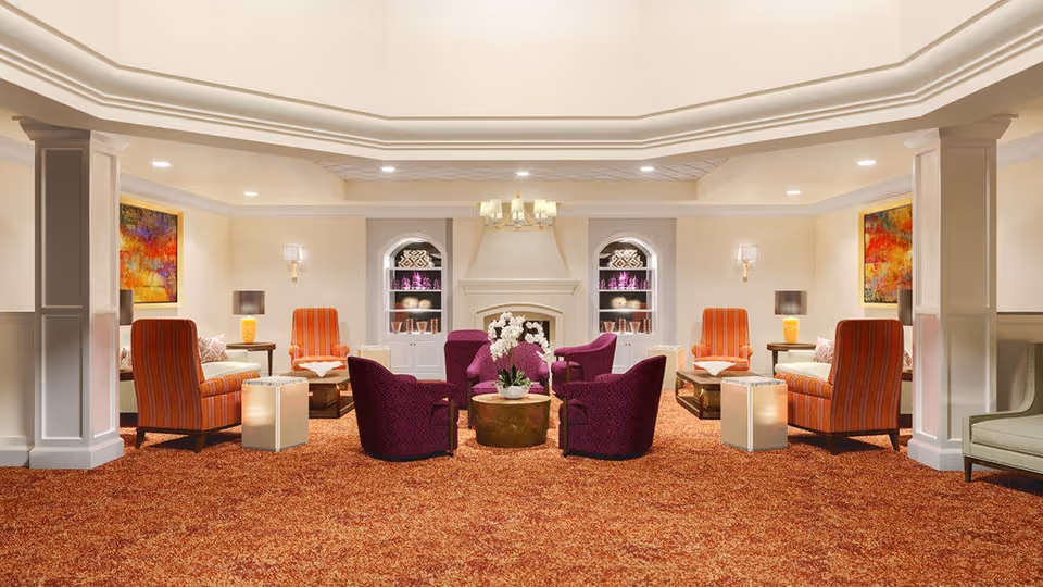 A spacious and elegant living room area with a warm color palette. The room features a patterned orange carpet, four purple armchairs arranged around a round coffee table with white flowers, and four orange-striped armchairs paired with side tables and lamps. There are two built-in shelves with decorative items flanking a central fireplace. The walls are adorned with colorful abstract paintings, and the ceiling has recessed lighting and a chandelier above the seating area.
