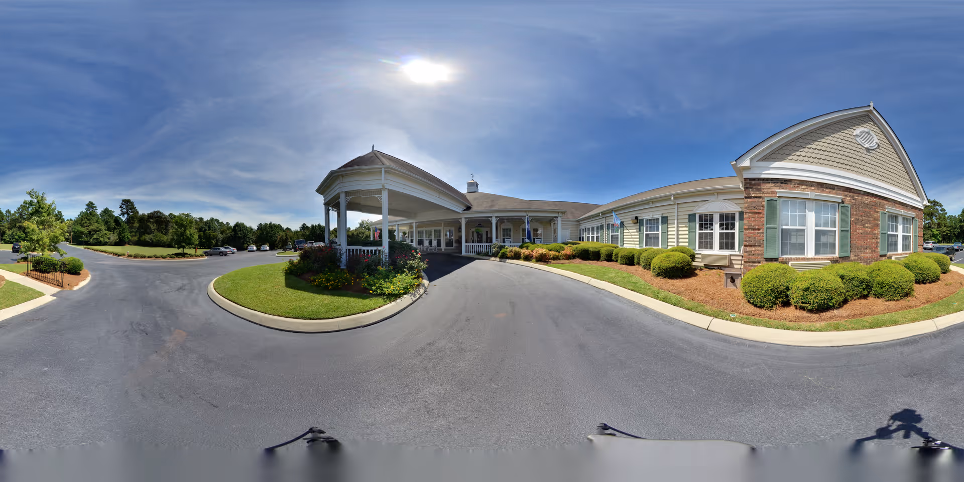 Wide panoramic exterior view of The Legacy of Camden senior living facility on a sunny day. The building features a covered entrance with white columns, beige siding, brick accents, and green shutters. Well-maintained landscaping with bushes and flowers surrounds the entrance. A paved driveway and parking area are visible, along with a clear blue sky and some trees in the background.