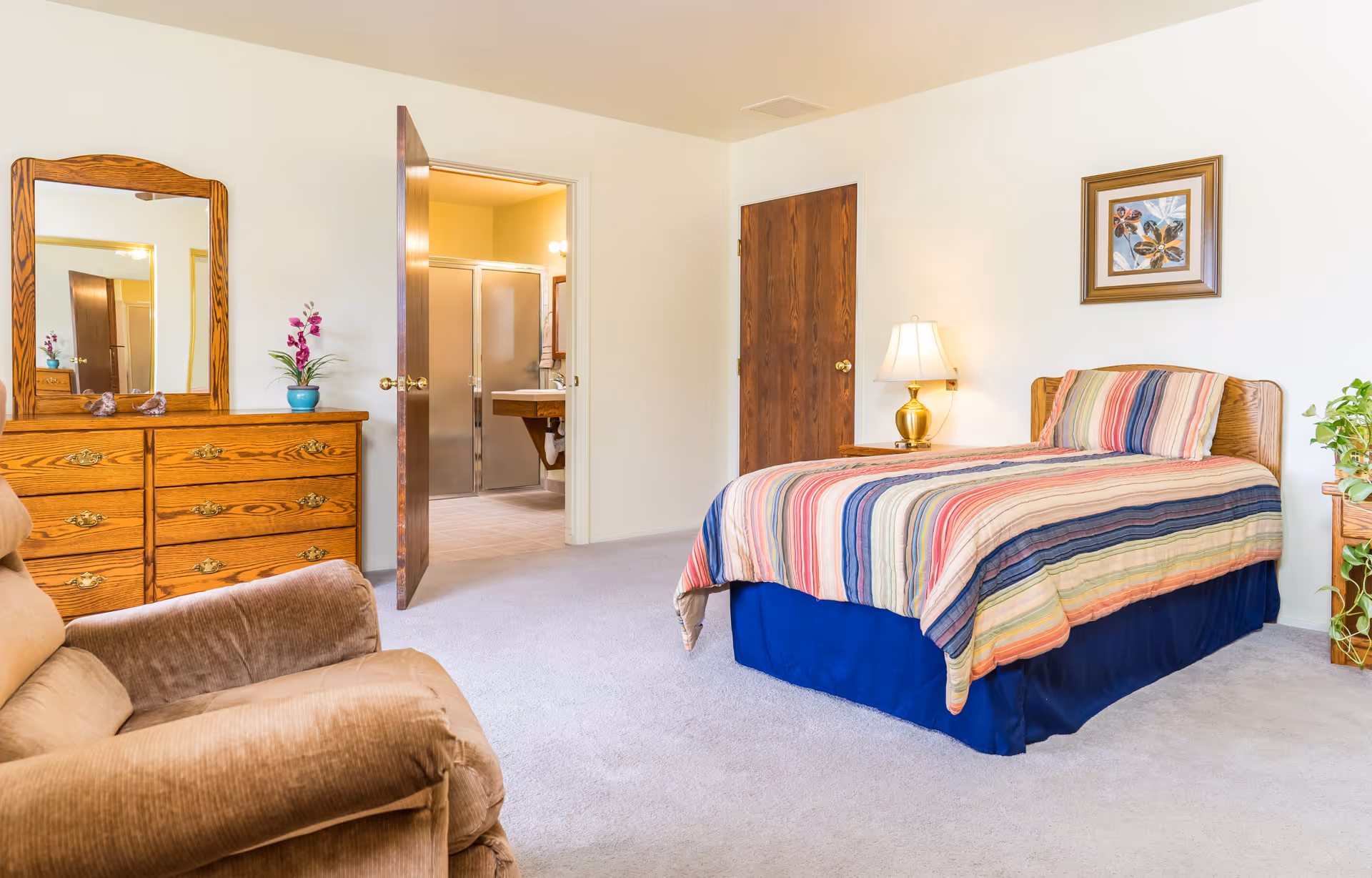 A bright assisted-living bedroom with a single bed with a striped bedspread, wooden dresser and mirror, recliner chair, and an open door leading to an en-suite bathroom.