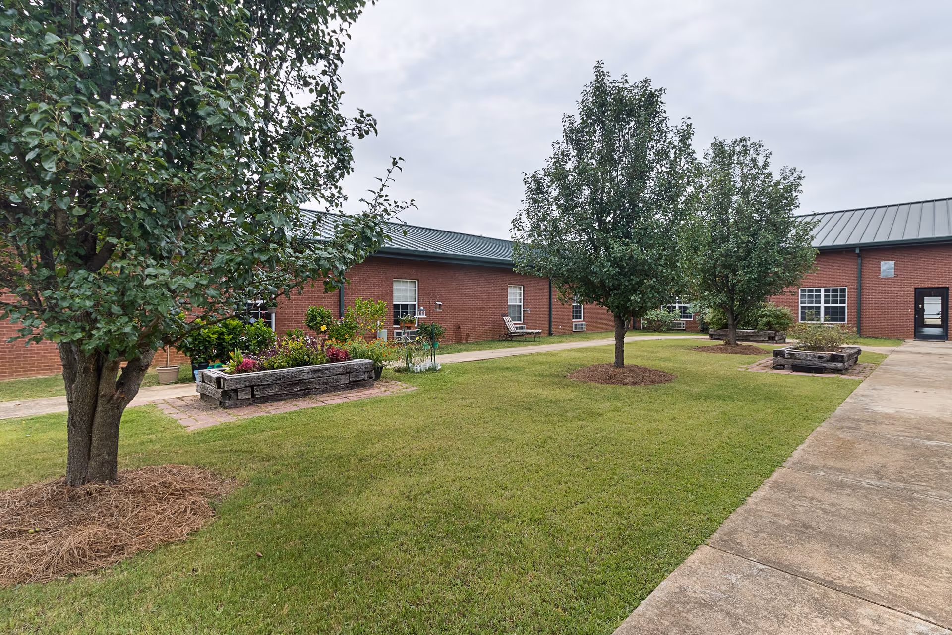 A grassy courtyard with trees, raised flower beds and walkways surrounded by a single-story brick assisted living building.