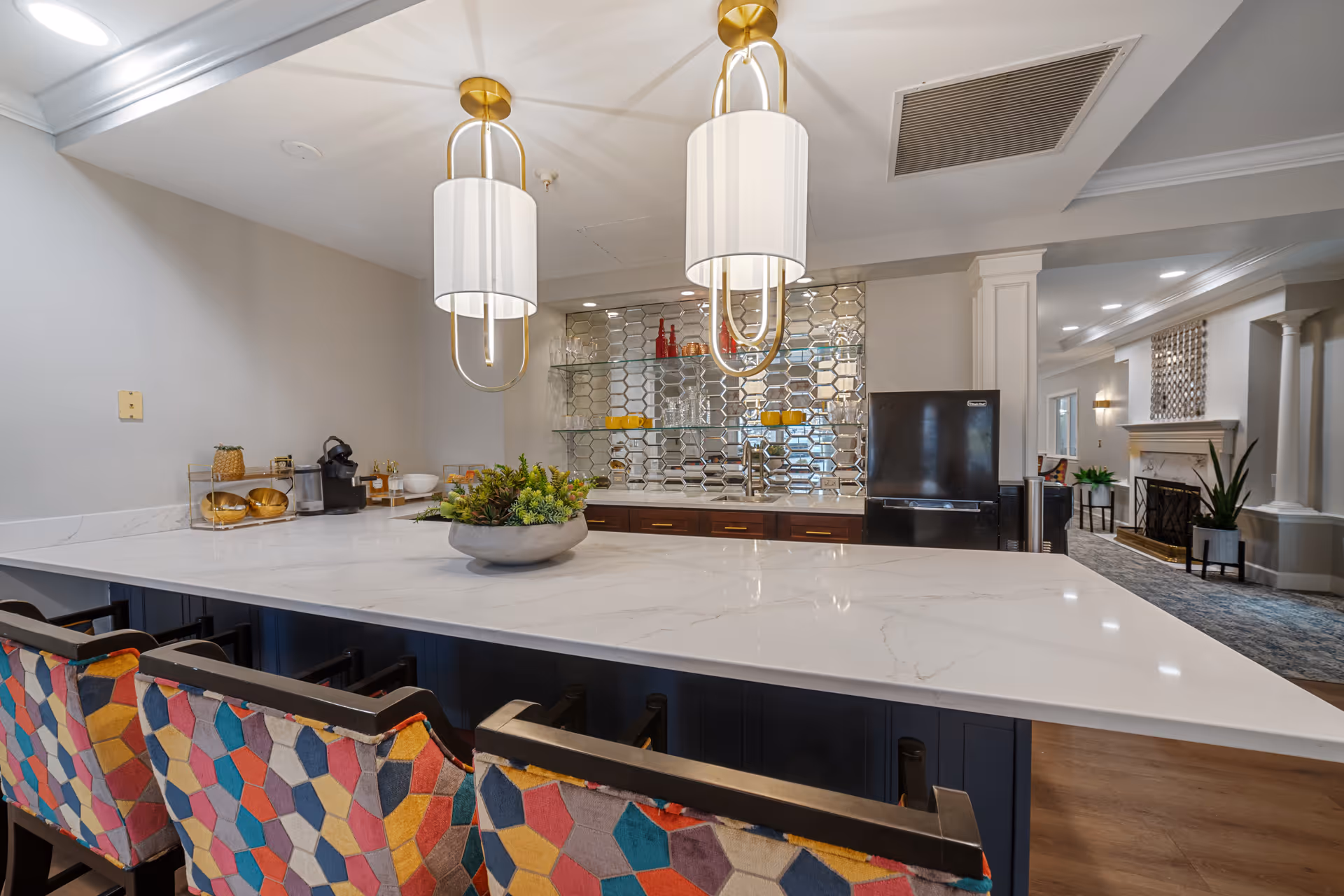 Modern kitchen area with a large white marble countertop island surrounded by colorful patterned chairs. Two elegant pendant lights hang above the island. The back wall features a mirrored backsplash with glass shelves holding glassware and decorative items. A black refrigerator is visible to the right, and a cozy living area with a fireplace and plants can be seen in the background.
