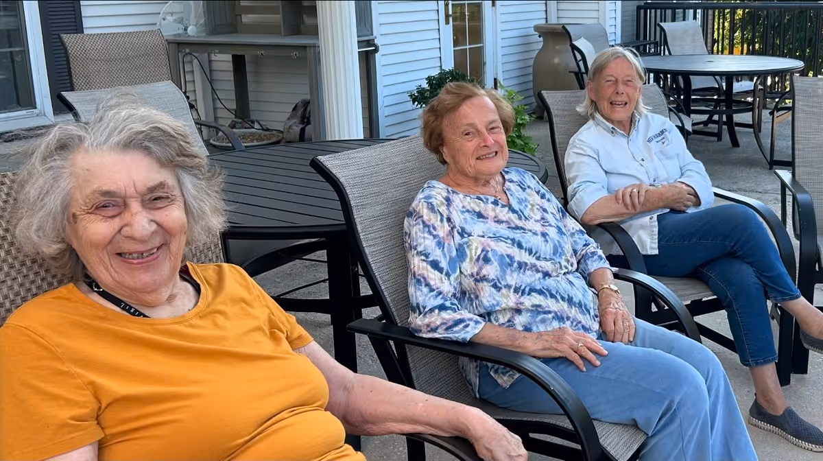 Three elderly women sitting and smiling on outdoor patio chairs at a senior living facility. They are seated around a table on a concrete patio with the building exterior visible behind them.