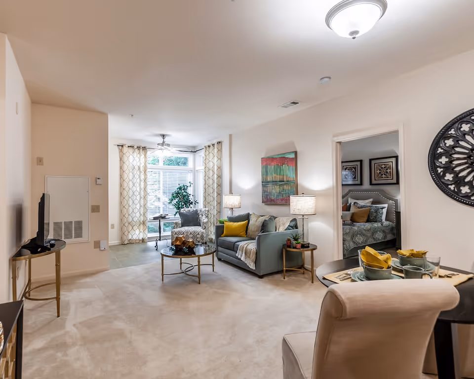 A cozy living room area with a light blue sofa adorned with yellow and gray pillows, a round glass coffee table, and two lamps on side tables. There is a patterned armchair near a window with sheer curtains, and a ceiling fan above. To the right, there is a small dining table set with plates, bowls, and yellow napkins. An open doorway reveals a bedroom with a bed and framed artwork on the walls. The walls are painted white and the floor is carpeted.