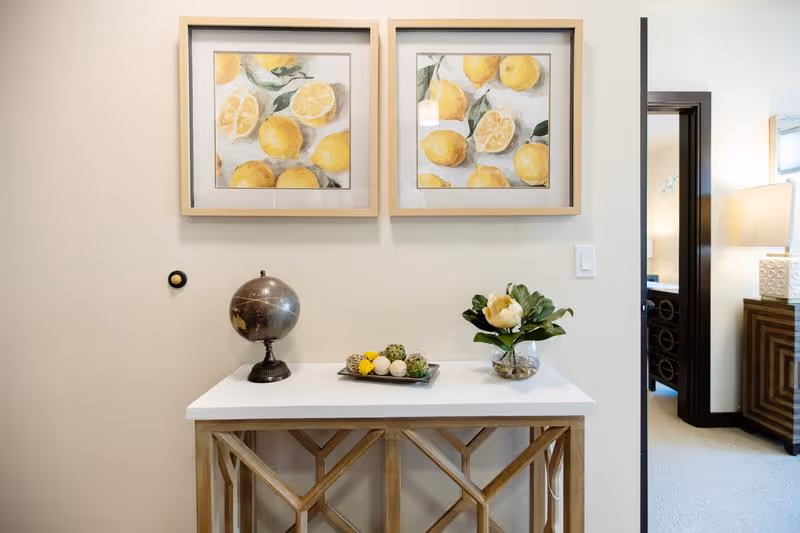 A decorative console table with a globe, a tray with decorative balls, and a vase with a flower. Above the table are two framed paintings of lemons. To the right, there is a doorway leading to a room with a lamp and furniture visible.