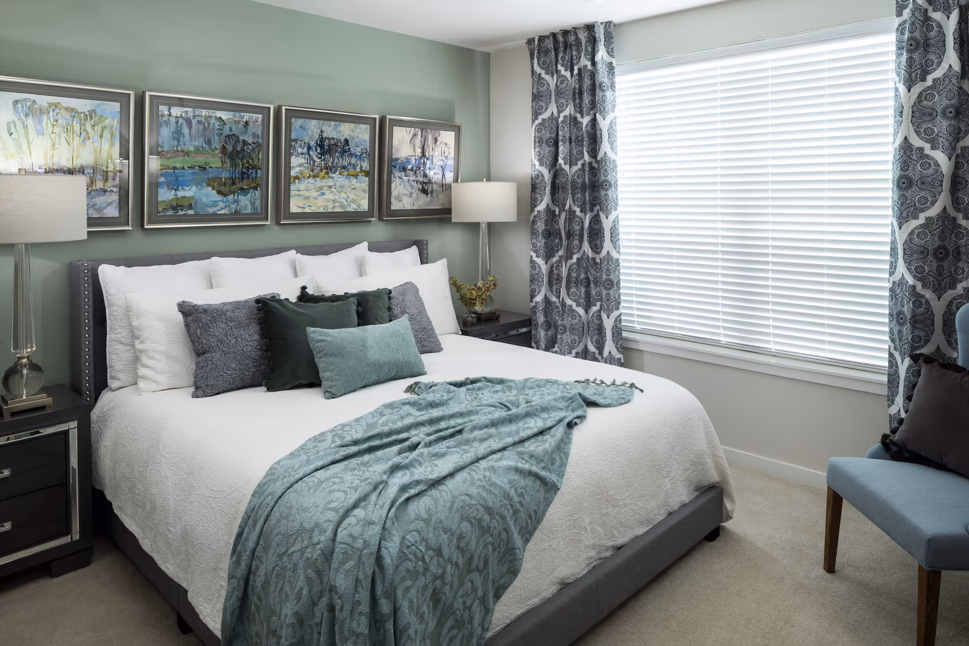 A neatly made bed with white bedding and multiple decorative pillows in shades of green and gray. A teal patterned throw blanket is draped across the foot of the bed. On either side of the bed are dark nightstands with glass lamps. Above the bed are four framed landscape paintings. To the right, a large window with white blinds and patterned curtains lets in natural light. A blue upholstered chair with a dark pillow is partially visible in the corner.