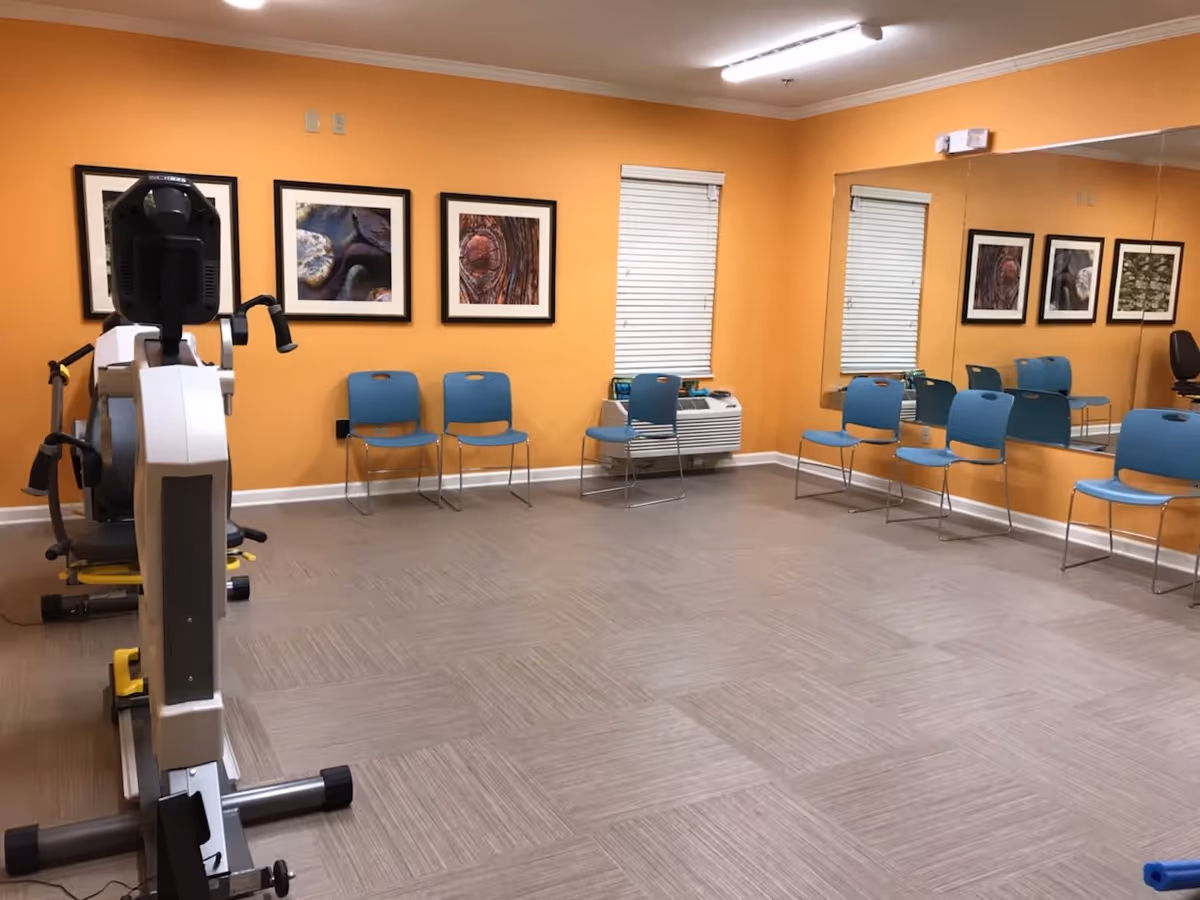 A fitness room with orange walls, a large mirror on one side, several blue chairs lined up against the walls, framed pictures hanging on the walls, and an exercise machine in the foreground.