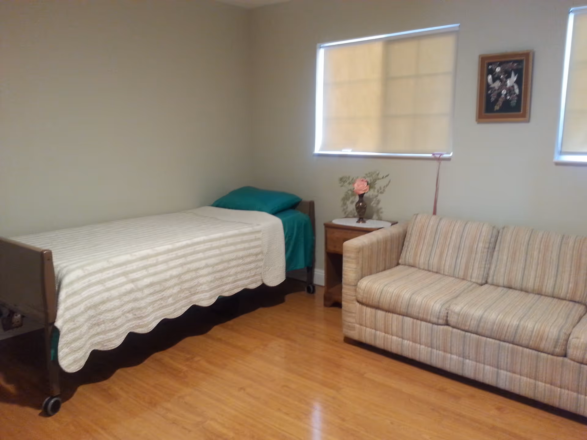 A simple room with a single bed covered with a white quilt and a green pillow, a wooden nightstand with a vase of flowers, a striped beige couch, two windows with beige blinds, and a framed picture on the wall.