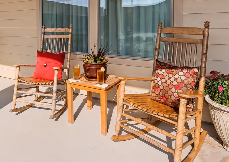 Two wooden rocking chairs with decorative cushions sit on a porch next to a small wooden table holding two glasses of iced tea and a flower pot. The porch is adjacent to a building with beige siding and large windows with vertical blinds. There is also a large planter with flowers on the right side.