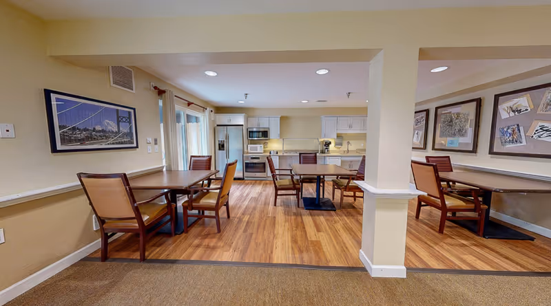 A spacious assisted living dining area with wooden floors and beige walls. The room features several wooden tables with cushioned chairs arranged around them. In the background, there is a kitchen area with white cabinets, a refrigerator, microwave, oven, and coffee maker. The walls are decorated with framed pictures and artwork. Recessed lighting illuminates the space.