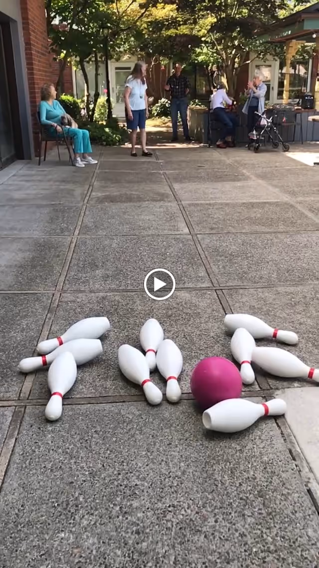 Outdoor patio area with a group of elderly people socializing near a building. In the foreground, several white bowling pins and a purple bowling ball are scattered on the concrete ground.