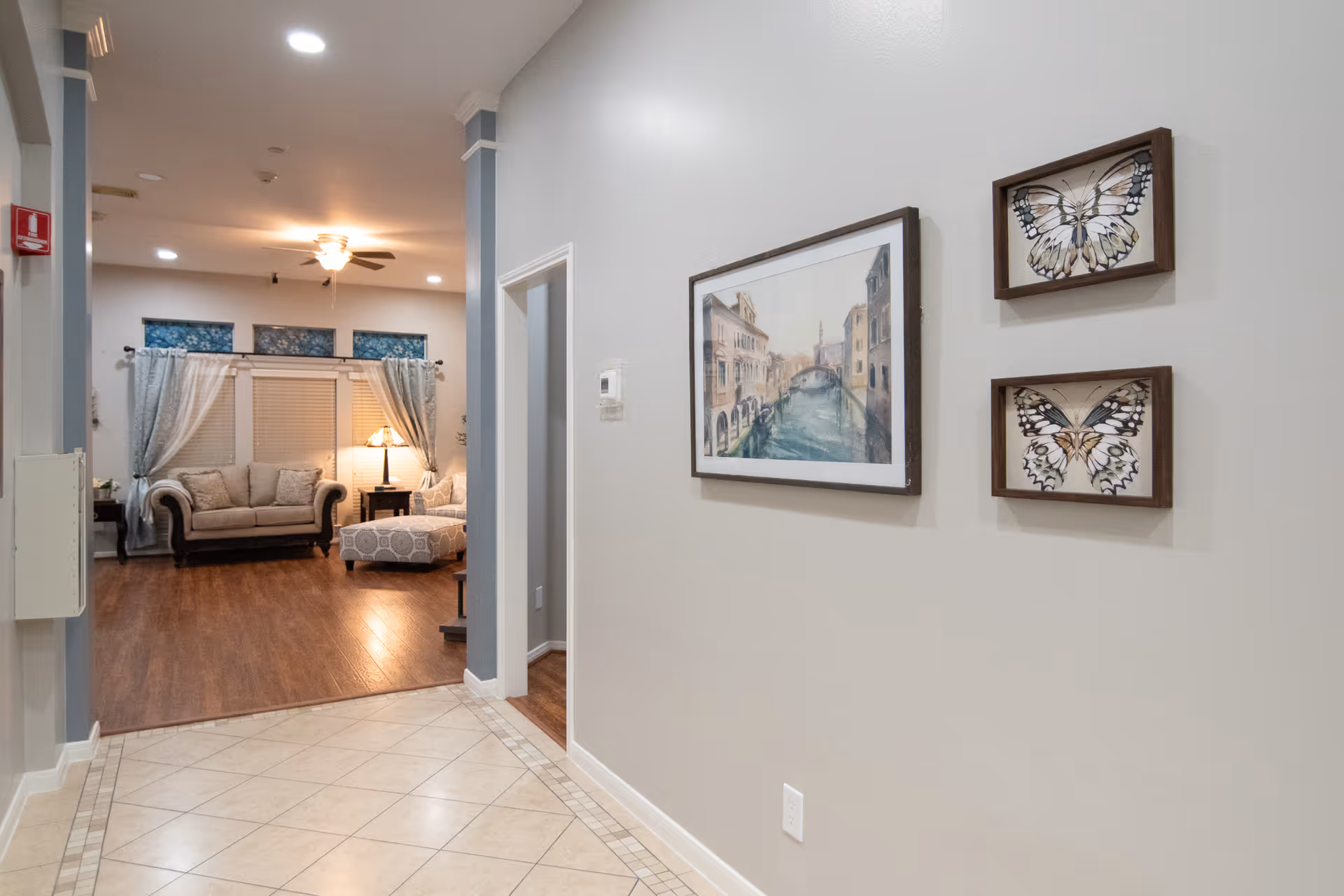 View down a hallway with tiled floor leading to a living room area with wooden flooring. The living room has a sofa, an ottoman, a side table with a lamp, and windows with curtains. On the right wall of the hallway, there are three framed pictures, one depicting a canal scene and two showing butterfly illustrations.