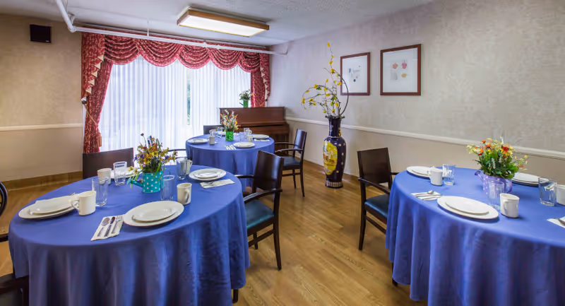 Dining room with three round tables covered with blue tablecloths, each set with white plates, cups, utensils, and glasses. Each table has a floral centerpiece. The room has wooden flooring, beige patterned wallpaper, a large window with sheer white curtains and red valances, a piano in the corner, and a tall decorative vase with yellow flowers.