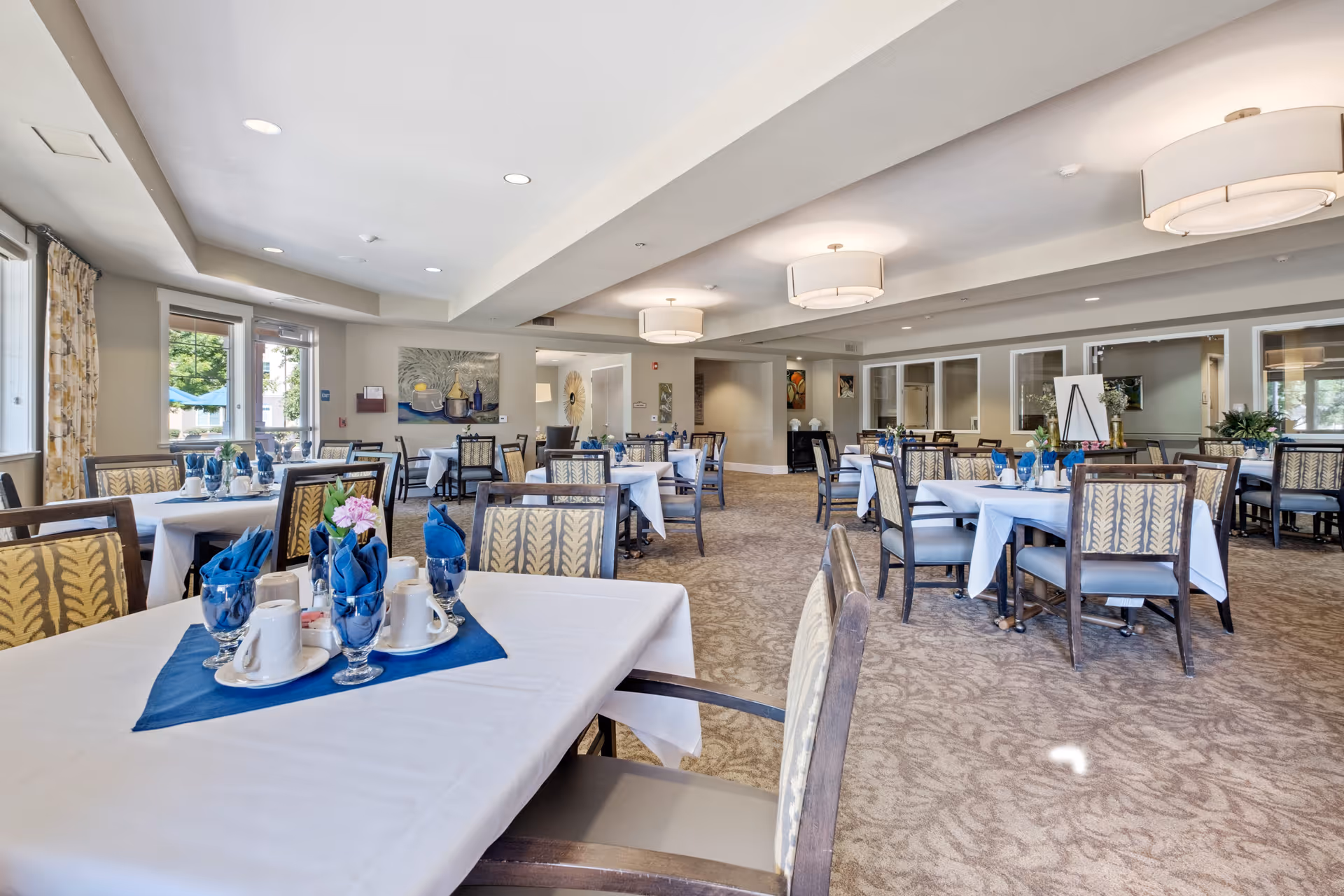 A spacious dining room in Carmel Village with multiple tables covered in white tablecloths and set with blue napkins, cups, and glasses. The room has patterned carpet, large windows with curtains, modern ceiling lights, and decorative artwork on the walls.