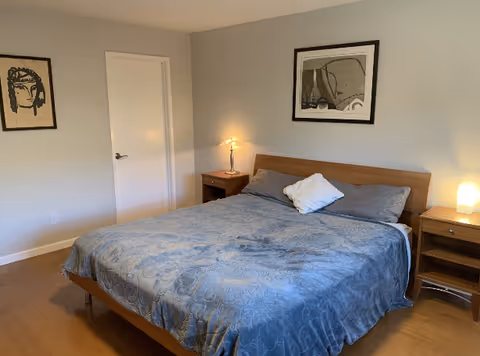 Bright bedroom featuring a large bed with a blue bedspread, two nightstands with lamps, and framed artwork on the walls.