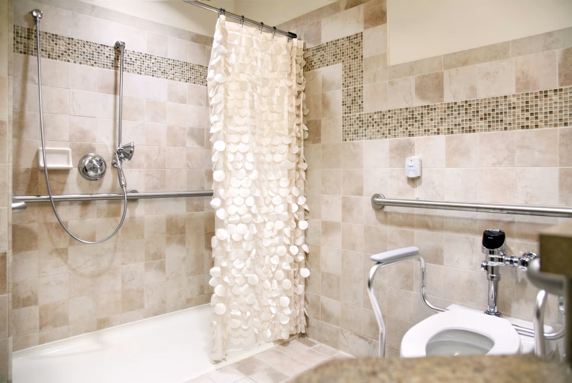 Accessible bathroom with beige tiled walls and floor, featuring a bathtub with a handheld showerhead and a white textured shower curtain. There are horizontal grab bars on the walls and a toilet with support rails.
