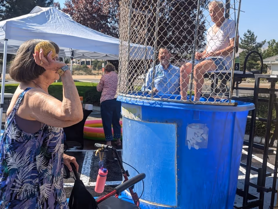 An older woman aims a ball at a dunk tank during an outdoor event where a seated senior waits inside the tank, with tents and onlookers in the background.