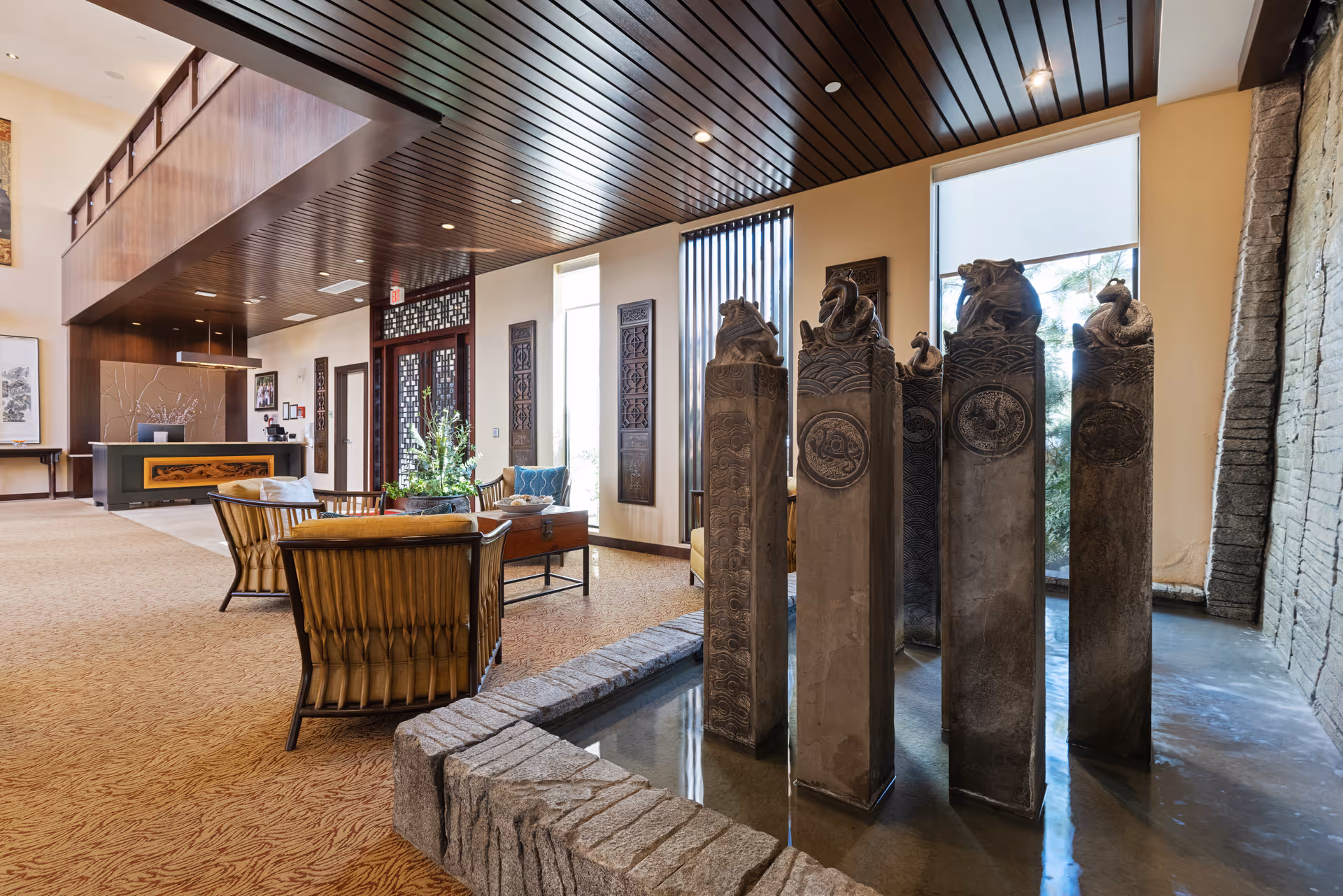 Interior view of a senior living facility lobby area with a decorative water feature consisting of tall stone pillars with carved animal figures on top. The space includes comfortable seating with wooden chairs and a table, large windows allowing natural light, and a wooden ceiling with recessed lighting. The reception desk is visible in the background.