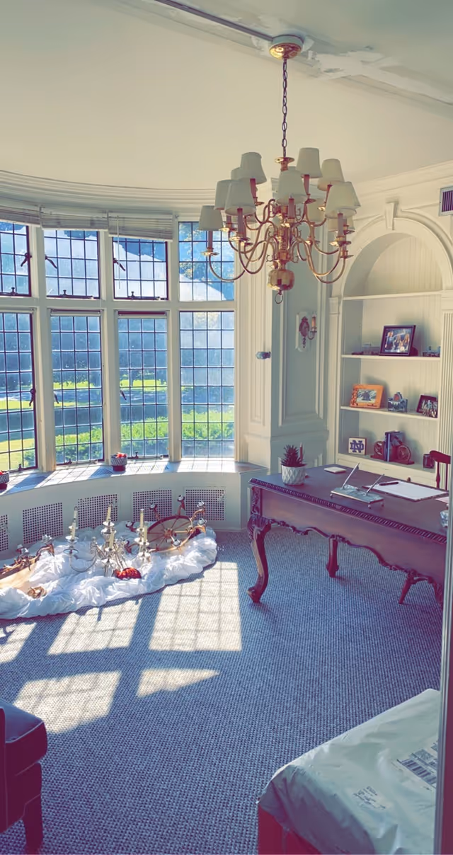 A bright room with large leaded glass windows letting in sunlight, a wooden desk with papers and a small plant on it, built-in shelves with framed photos and decorative items, a chandelier hanging from the ceiling, and several chandeliers placed on a white cloth on the carpeted floor.