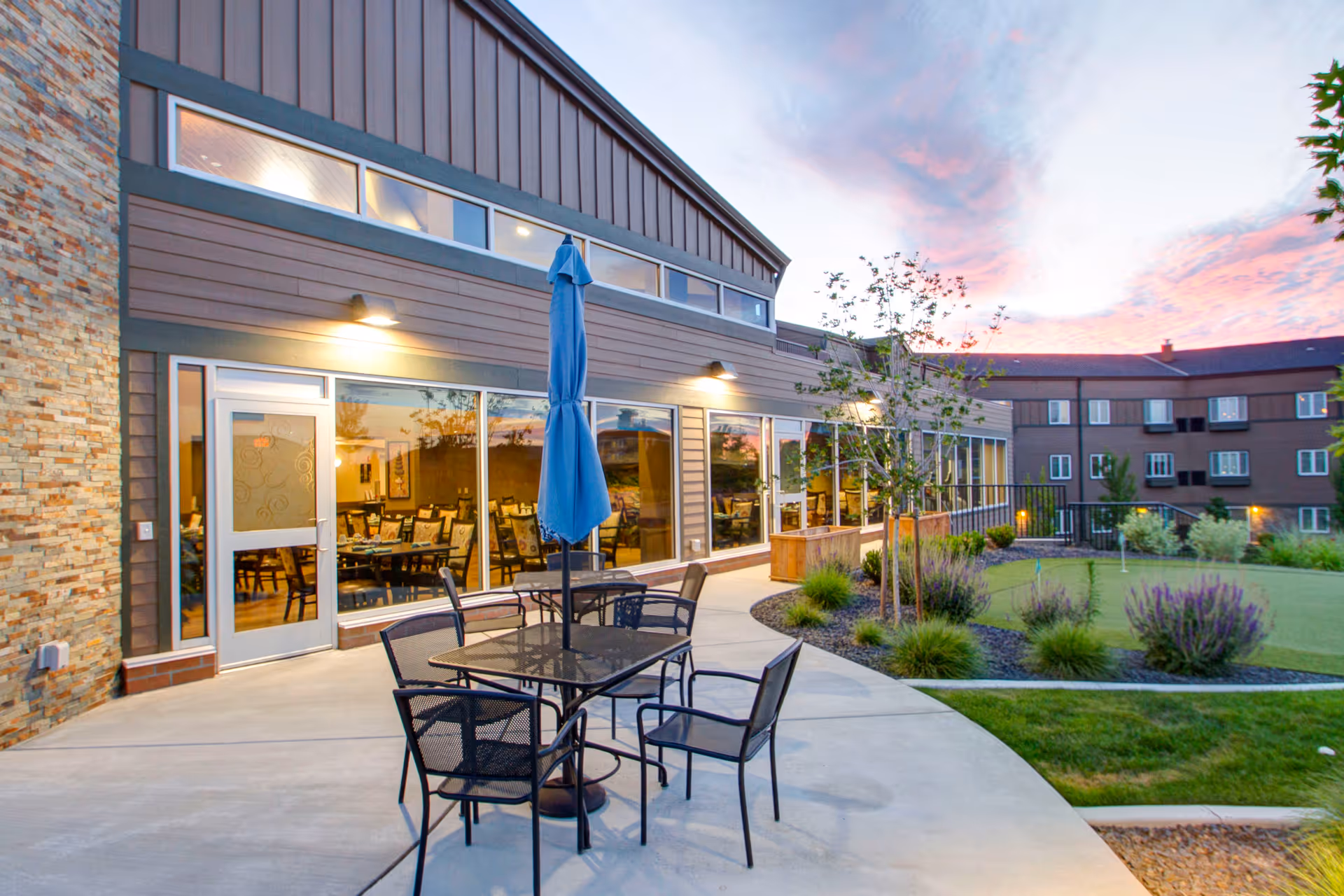Outdoor patio area at a senior living facility with a glass table and four metal chairs under a closed blue umbrella. The patio is adjacent to a building with large windows showing a dining room inside. The area includes landscaped greenery and a putting green, with a multi-story residential building in the background under a colorful sunset sky.