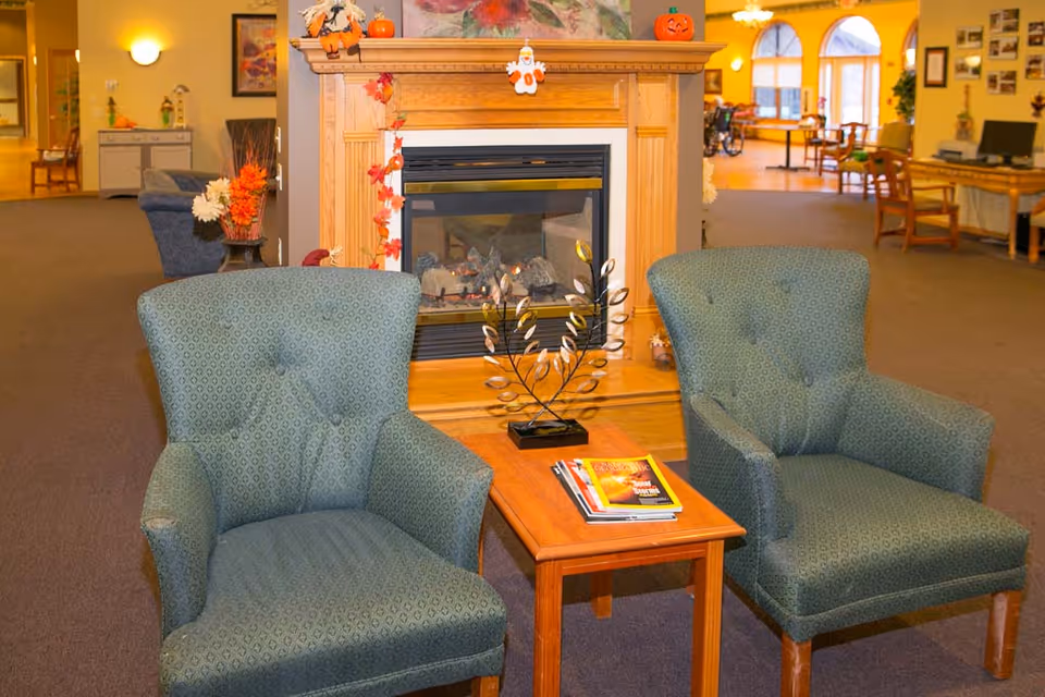 A cozy senior living facility common area with two green upholstered armchairs facing a wooden coffee table that holds magazines and a decorative metal sculpture. Behind the chairs is a wooden fireplace mantel decorated with small Halloween-themed items including a pumpkin and a ghost. The room has warm lighting and in the background, there are more seating areas and large windows letting in natural light.