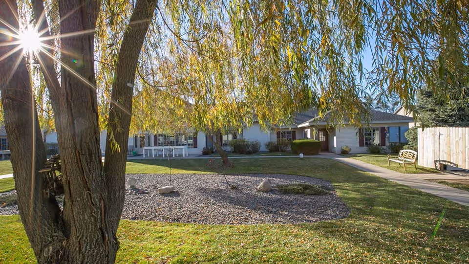 A sunny outdoor view of a senior living facility with a large tree in the foreground, a landscaped rock garden, green grass, a paved walkway, benches, and a single-story building with white siding and maroon shutters in the background.
