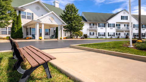 Wooden bench on a lawn beside a sidewalk leading to the covered entrance of a two-story white building with a green roof.